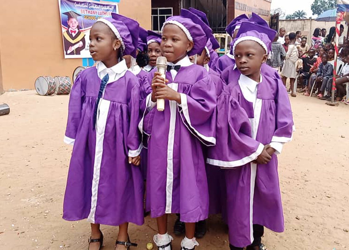 Young pupils in purple graduation gowns and caps at an outdoor ceremony
